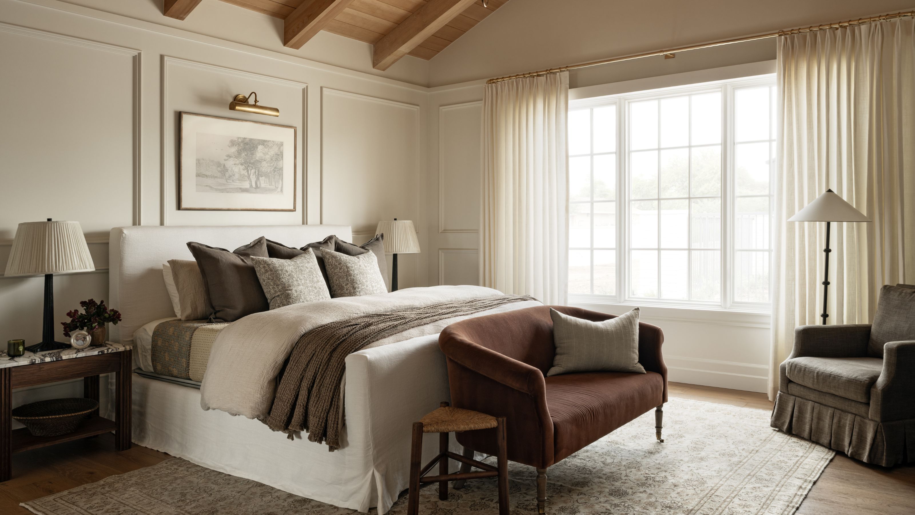 a calm but large primary bedroom with a vaulted oak ceiling, paneled walls, large windows with linen drapes, a white linen bed with a burgundy settee and a small skirted arm chair