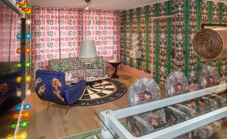 Always Close installation by Studio Job in Luxembourg. A lounge area with a leopard print sofa, a blue chair, an oval glass coffee table, a tree shaped side table, a dartboard patterned rug, a glass dining table with chairs and colourful patterned wall paper.