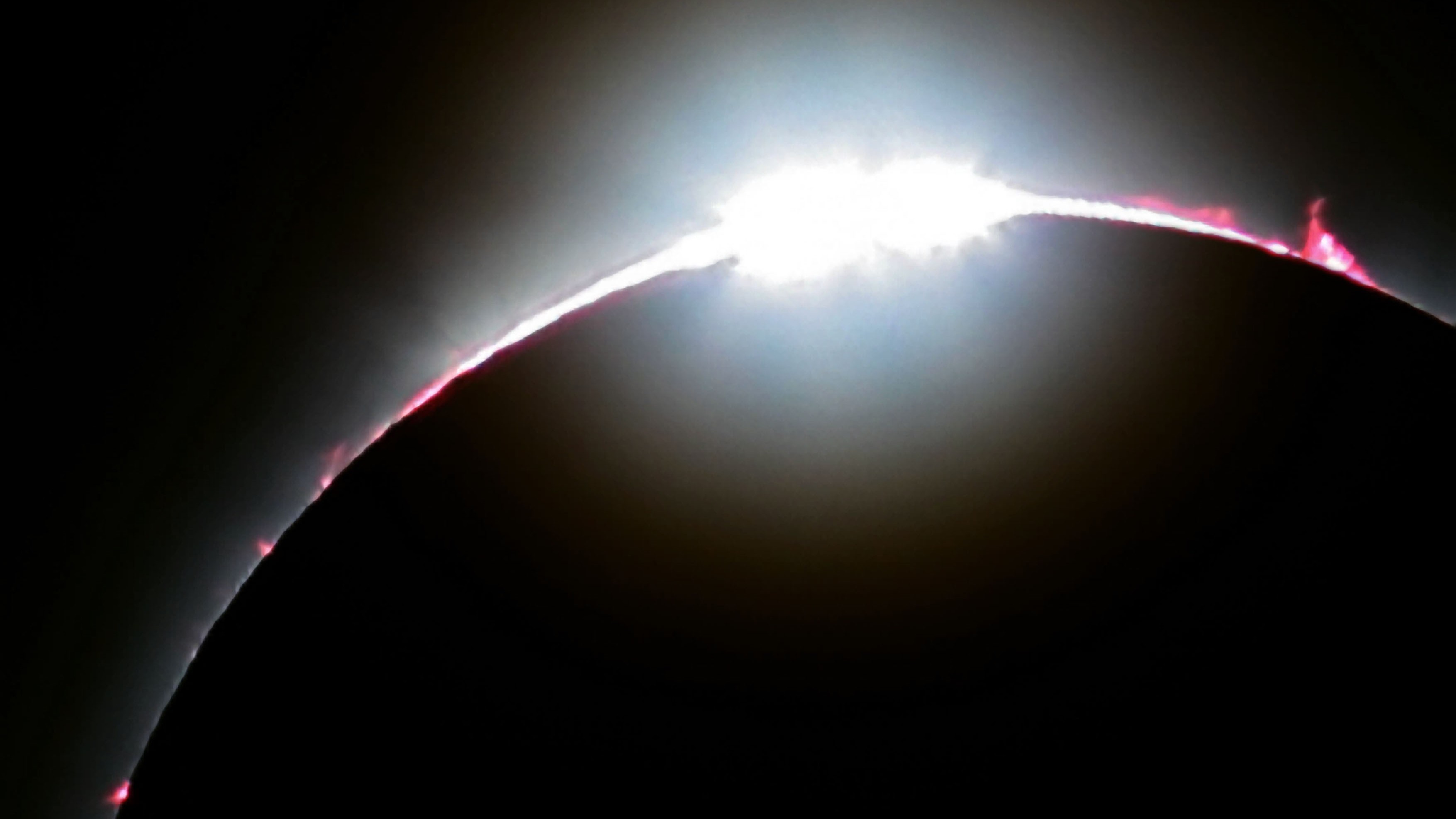 a bright light shines from behind the moon creating a diamond ring effect. there are bright pink prominences lifting off from the edge of the sun.