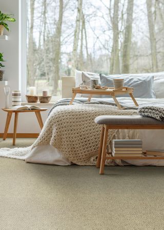 A bedroom with cream colored carpeting. On the carpet is a double bed with a food table on top of it. There is a side table next to the bed with a vase.