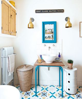 White bathroom with colourful patterned floor, a blue painted mirror on the wall and table made into a vanity