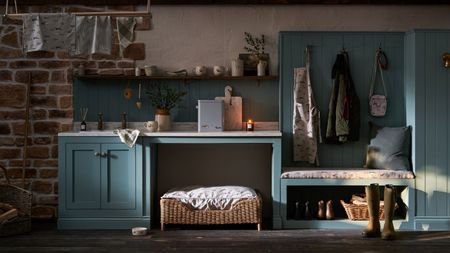 A blue boot room with exposed brick walls, brown wooden flooring, shelving, a dog bed, coats hanging up, green wellies, a lit candle, and laundry hanging up high to the top left.