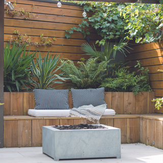 Tropical plants surround a patio, upon which can be found a bench and a firepit