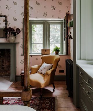 A cottage style bedroom with pink wallpaper and a yellow wingback armchair. Green curtains frame the windows, and a wooden four poster bed sites in front of an original fireplace