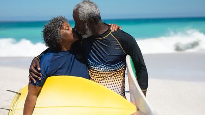 A happy retired couple kiss on the beach while holding surfboards.