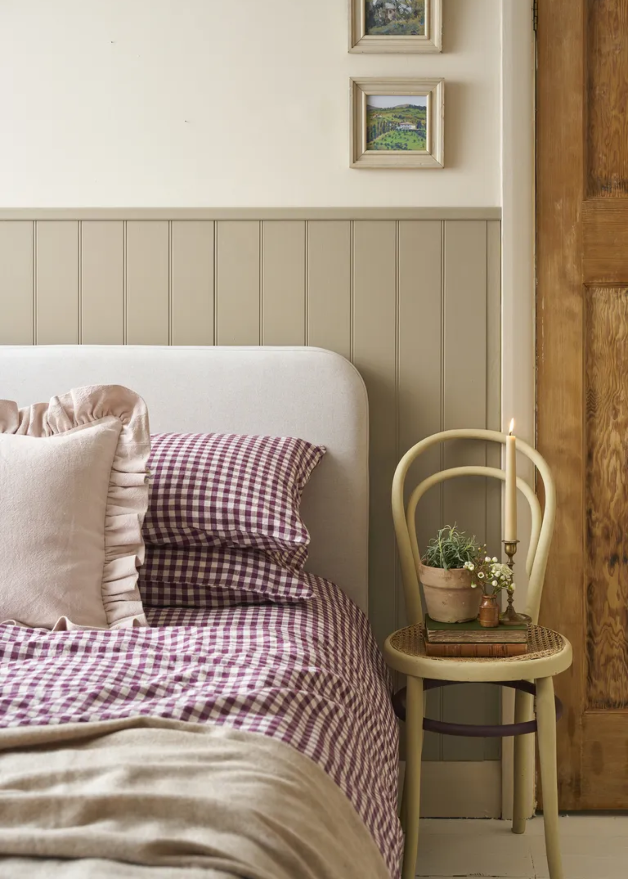 A cozy cottage style bedroom with a gingham bedscape and a chair with a potted plant, a mini vase, a taper candle, and a book