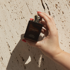 Hand with red nail polish holding up jo malone london cologne intense against beige stone wall 