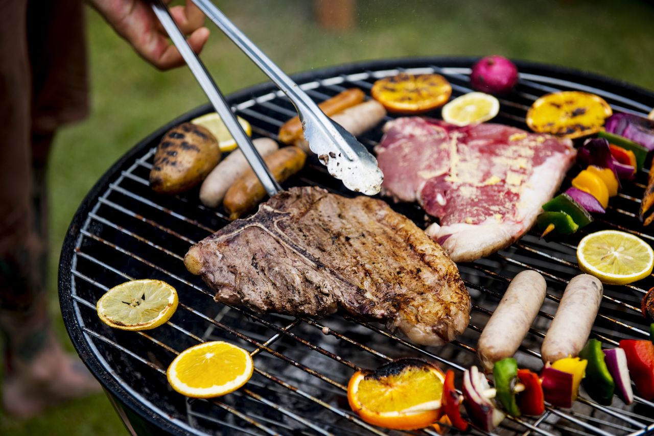 A closeup image of a steaks, sausages and slices of orange on a charcoal BBQ grill.