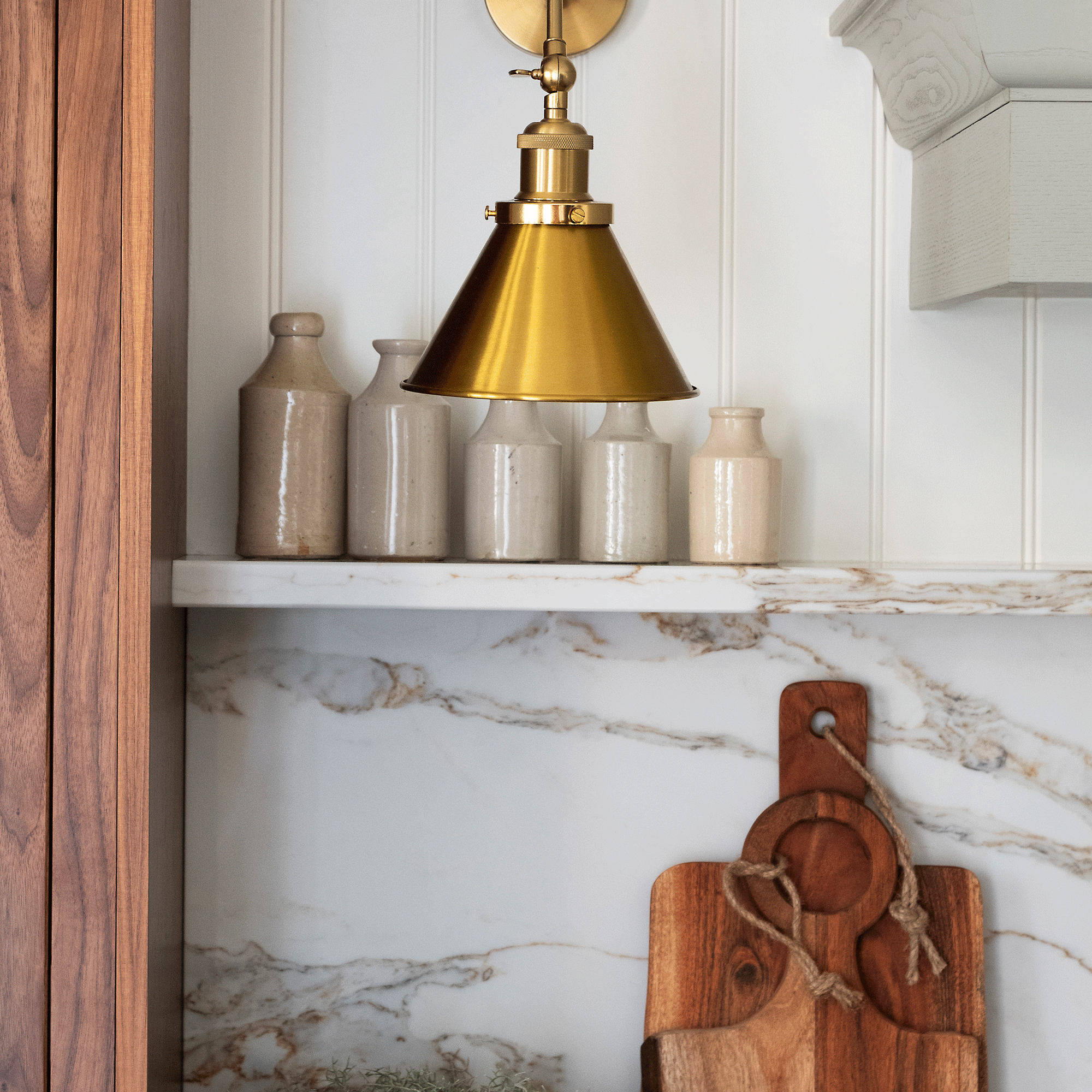 a neutral country-style kitchen with a marble splashback and a polished brass wall light and dark wood chopping boards