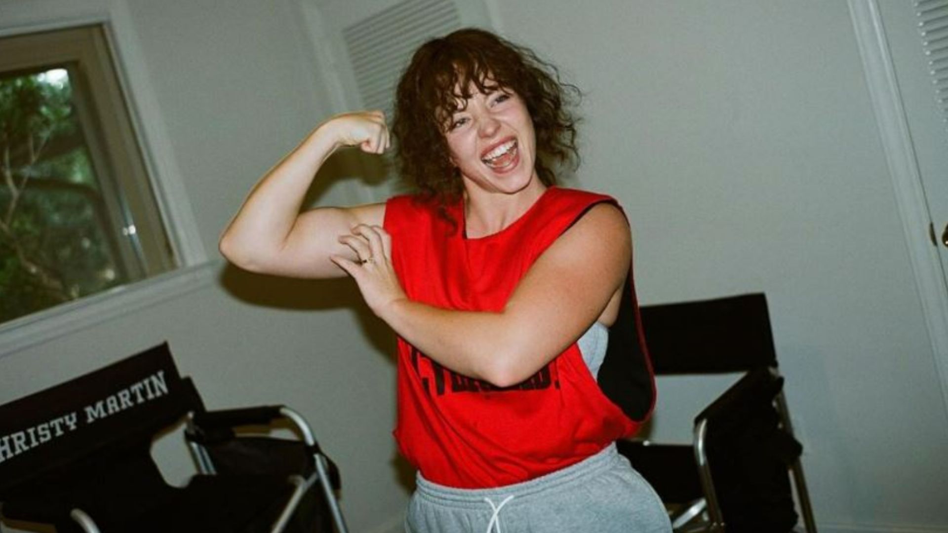 an image Sydney Sweeney posted on her instagram of her in character as Christy Martin, she is wearing a brown wig and red tank top, flexing her right bicep