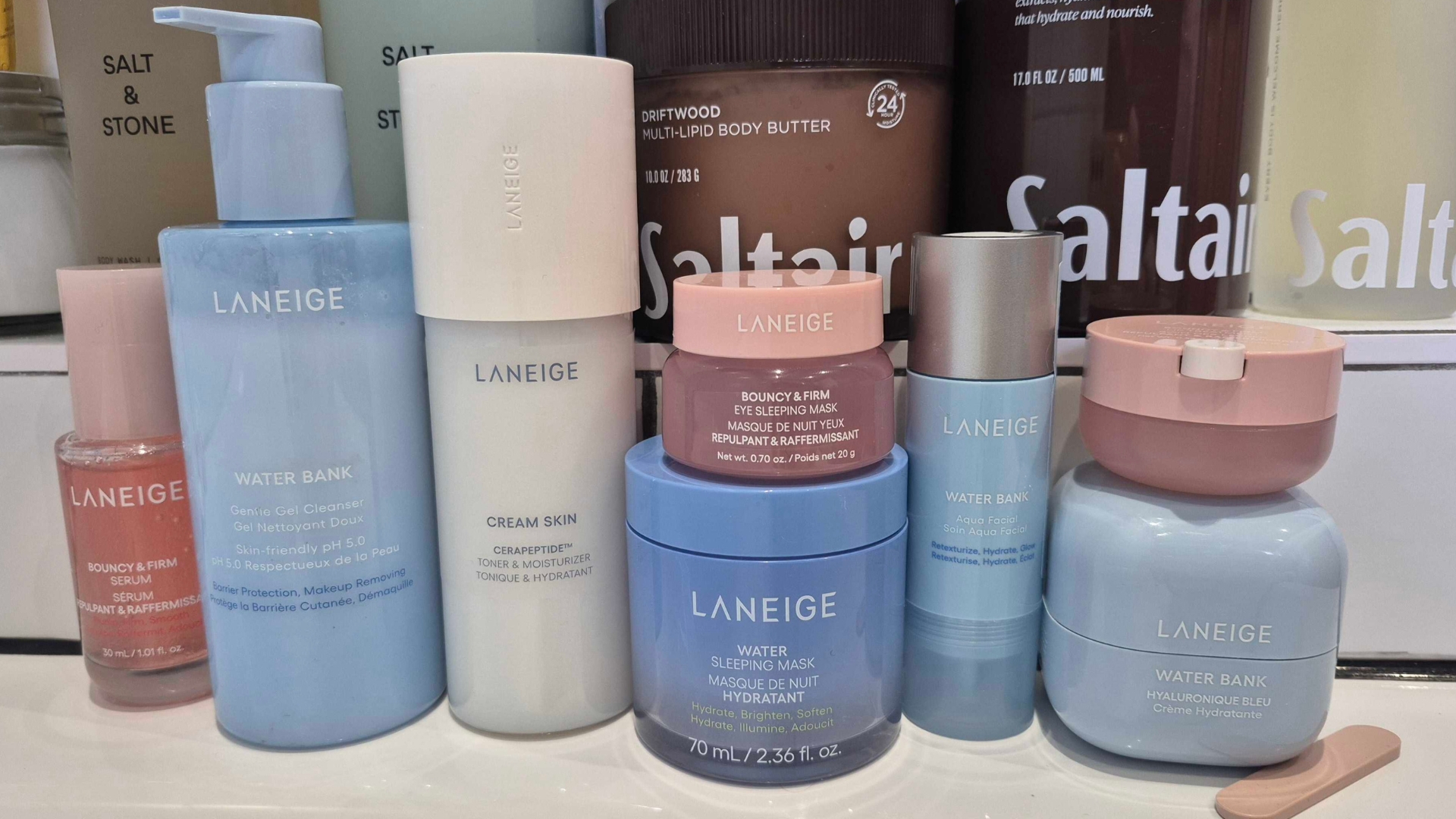 A picture of 8 Laneige products, 6 of which are featured in this 'best' guide, including the Water Bank Cleanser and Sleeping Eye Mask, all arranged in a line on the edge of a bath tub, surrounded by more skin and body products.