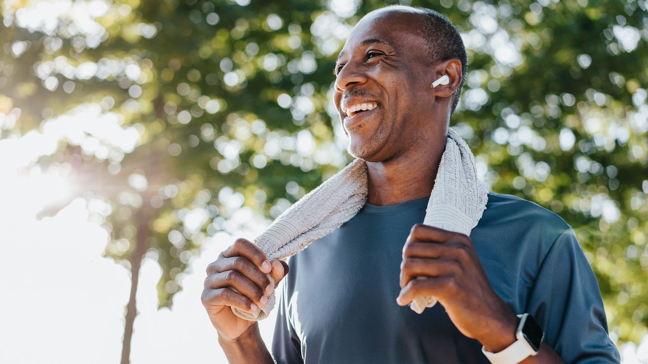 A smiling man stands outside, holding the ends of a sweat towel that is looped around his neck. He&#039;s wearing a light t-shirt and has earbuds in; he also has a sports watch strapped on his wrist.