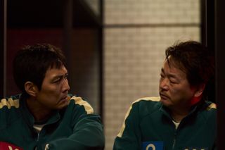 Two Korean men wearing matching tracksuits look at each other while sitting in a room with white brick walls, in 'Squid Game' season 2.