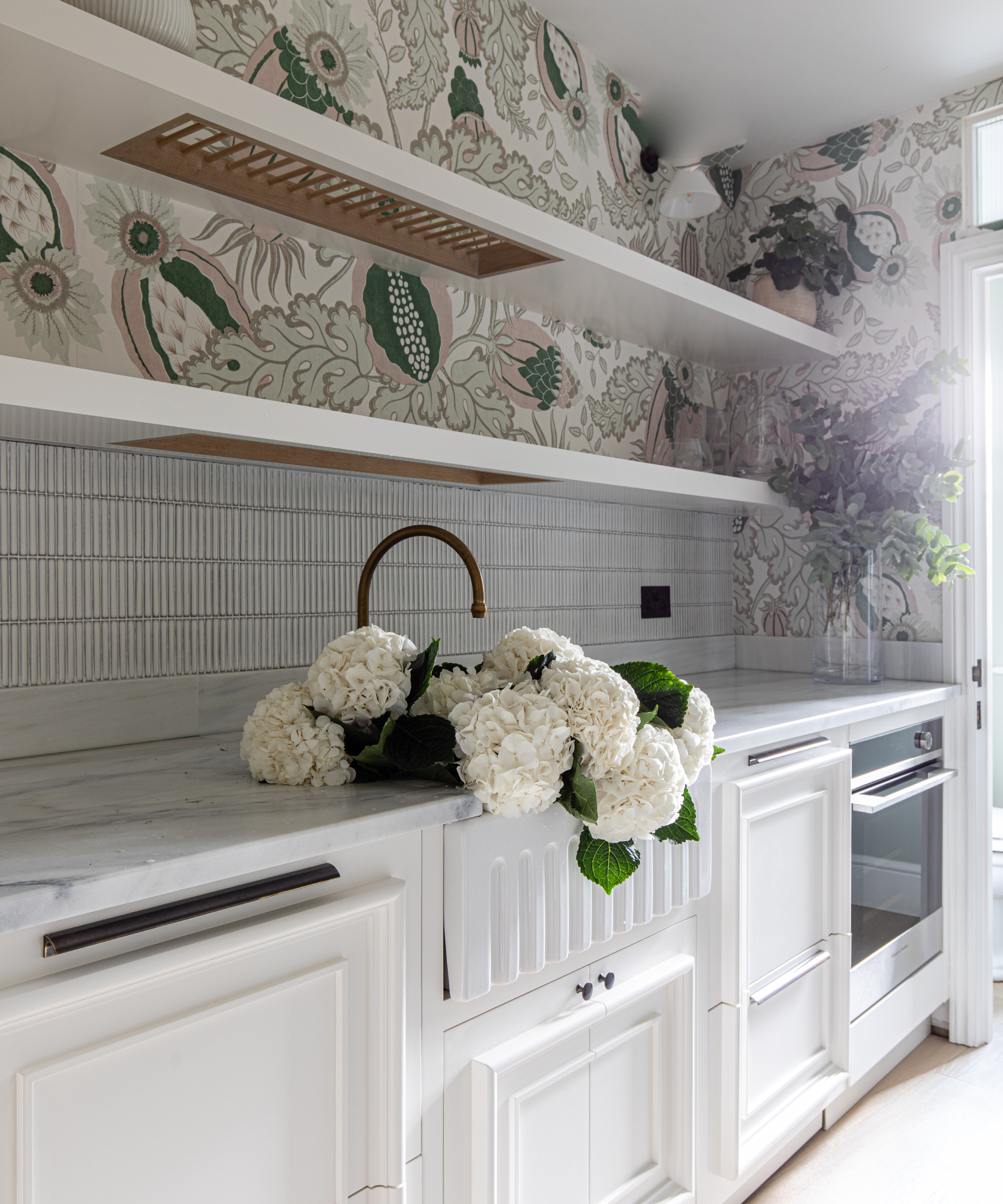 utility room back kitchen area with white units, white marble work top, white tiled splashback with large floral patterned wallpaper above, white shelves with insert in centre with rack, white flowers in sink