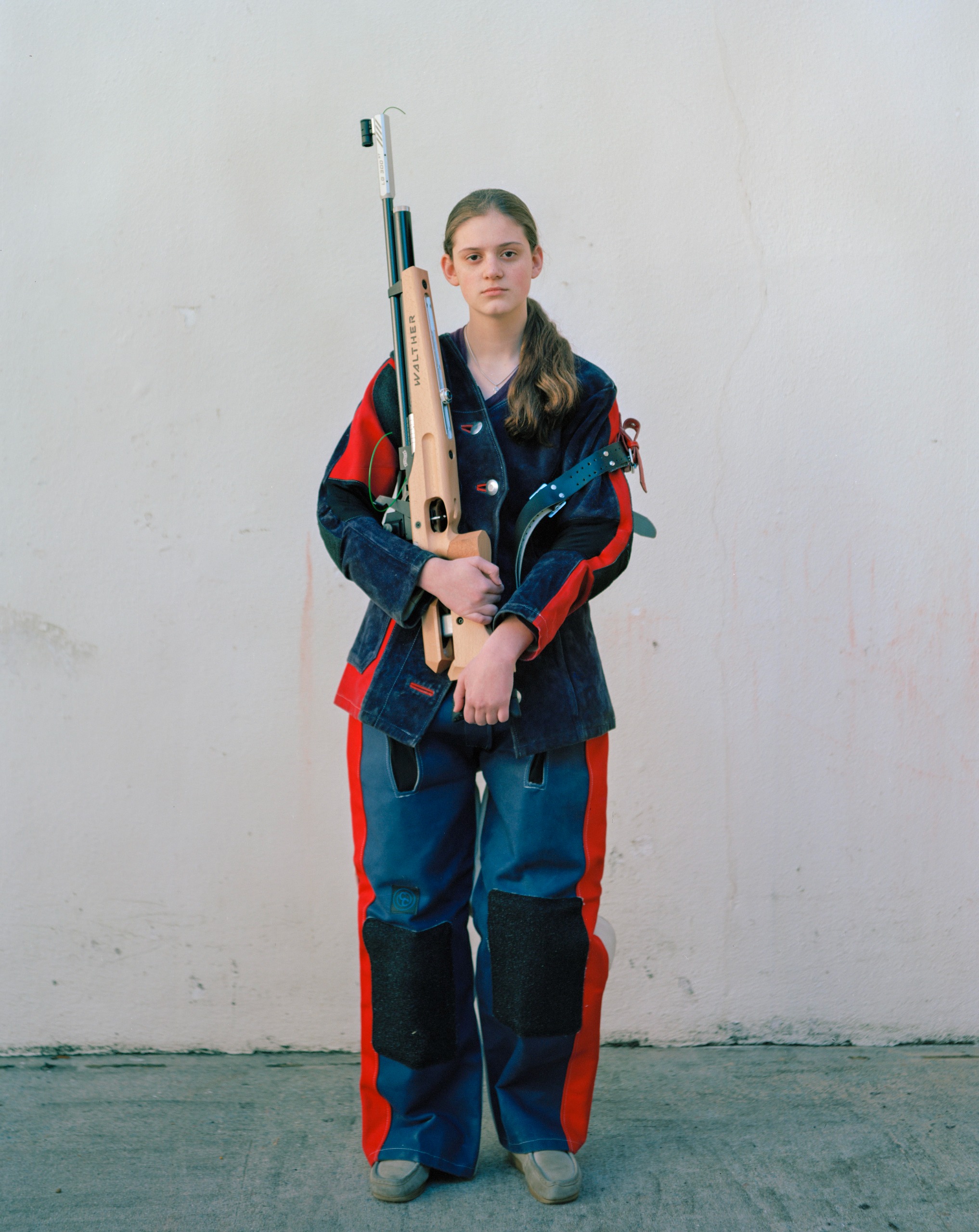 A teenage girl in a padded red and blue shooting suit holds a competition rifle upright against a white wall
