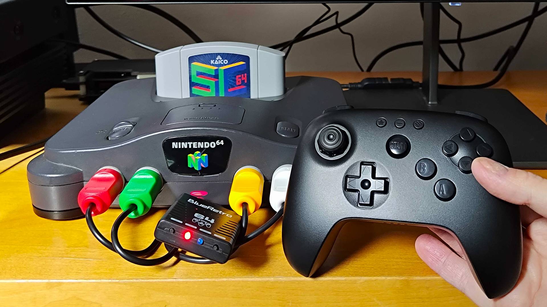 Hand holding 8Bitdo N64 controller next to original console with Blueretro Bluetooth receiver connected. 