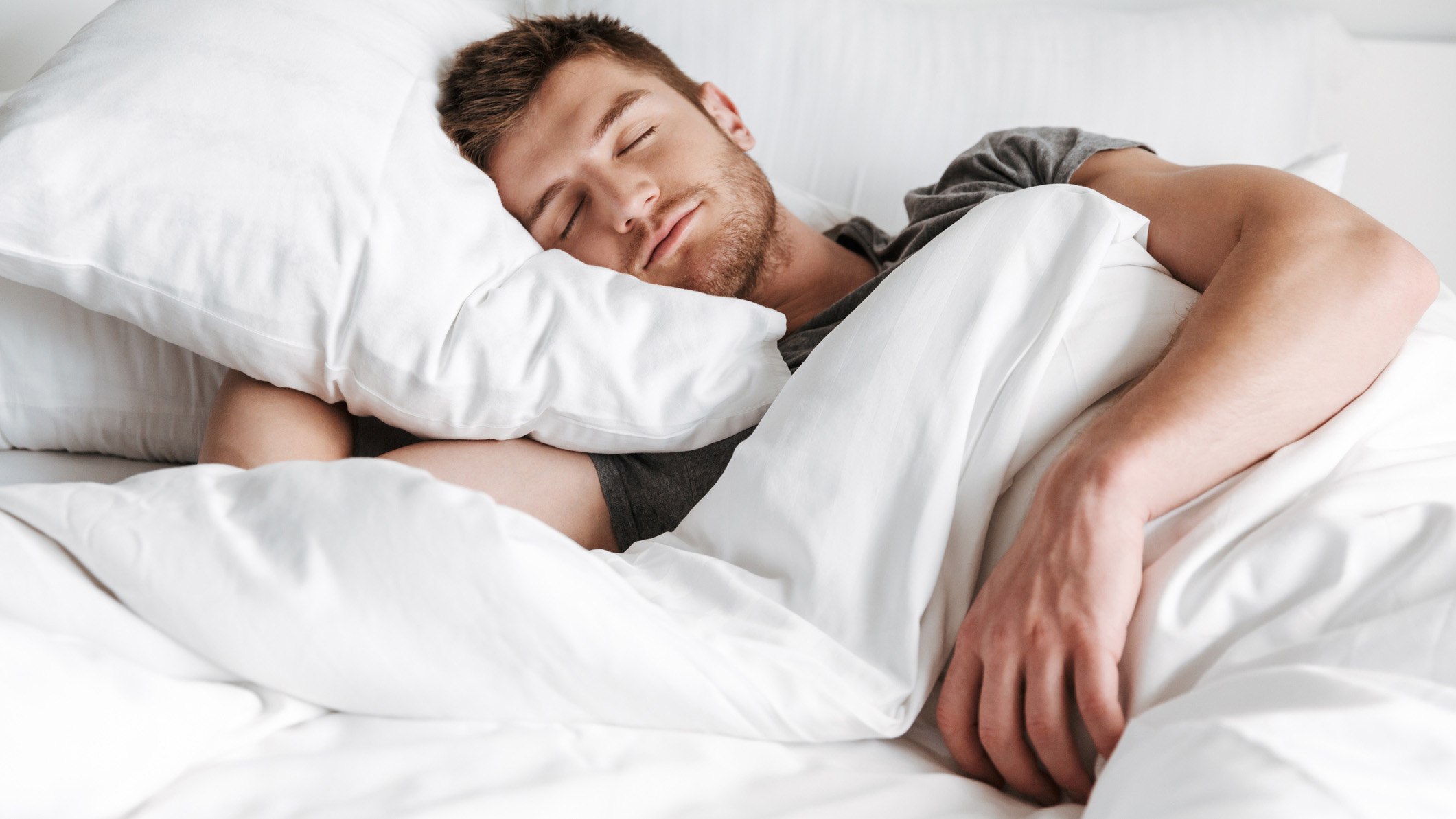 A dark-haired man is sleeping on his side, covered with a white duvet