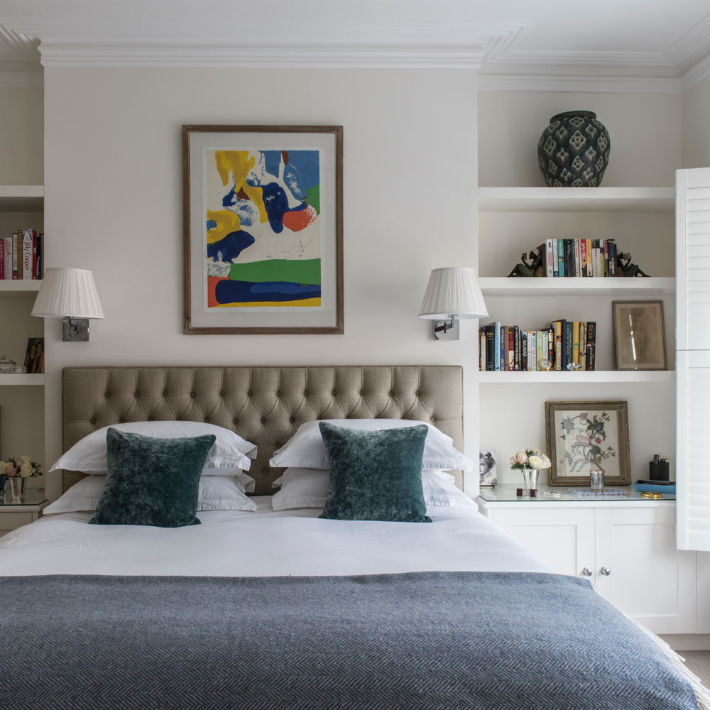 Small bedroom with double bed, headboard, wall lamps and shelving alcoves.