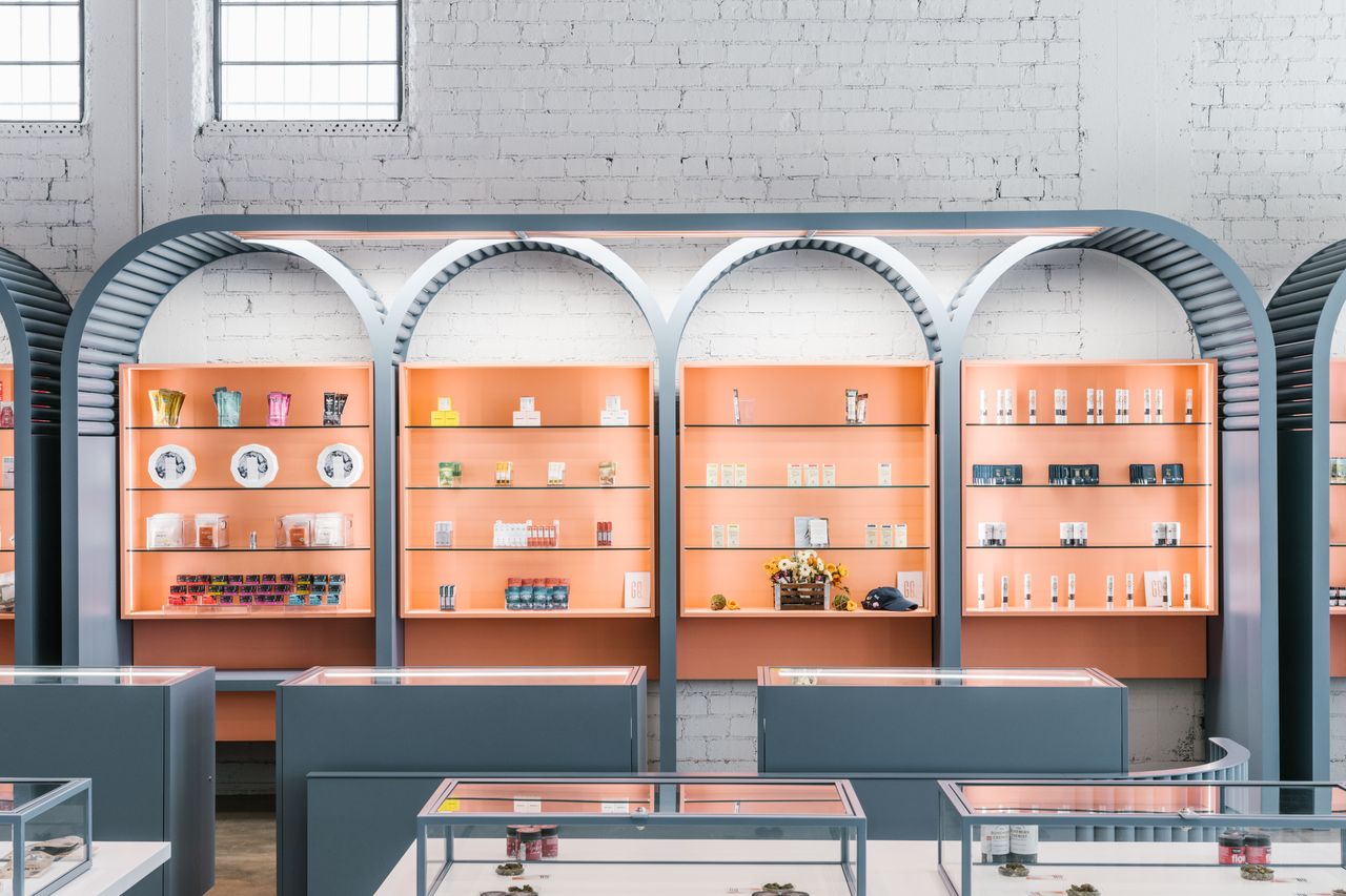 Green Qween Marijuana dispensary dispensary, Los Angeles. Grey counters with glass tops in front of an orange wall with shelves which are curved on the tops.