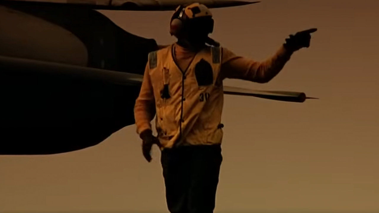 A man in a yellow very pointing to the left with a jet fighter behind him in Top Gun