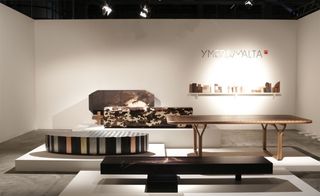 A room with a white shelf on a white wall stacked with books. A brown long bare dinner table. A camouflage-looking rectangular box placed on a white platform. a round black platform with colourful lines. Thick brown table with uneven legs
