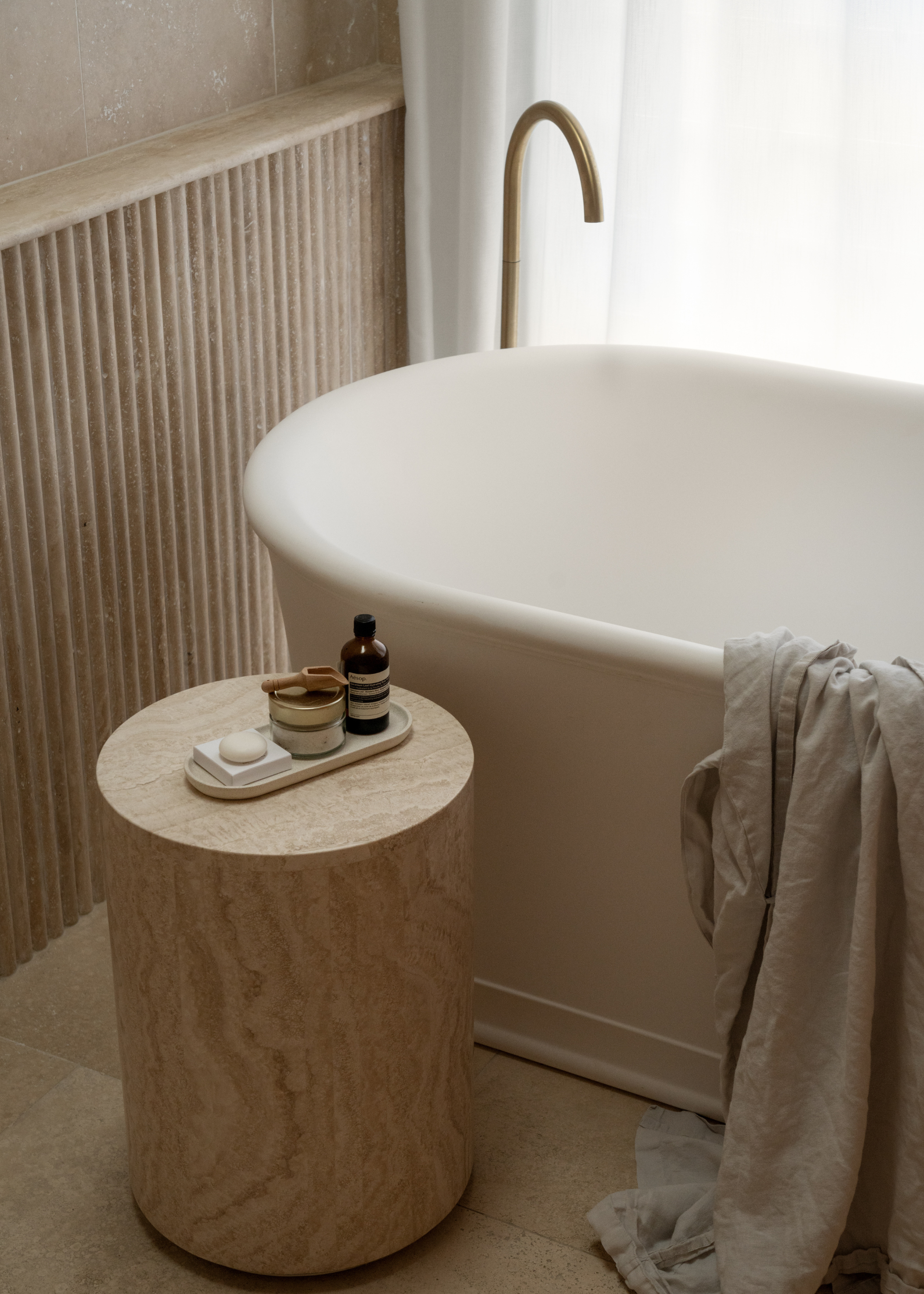 An airy bathroom with a standing tub, a bunched shirt, a wood stool with an Aesop bottle, a jar of bath salts, a bar of soap
