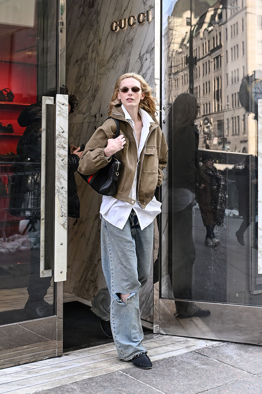 Alex Consani wearing a brown jacket, white button-down, blue jeans, and black loafers
