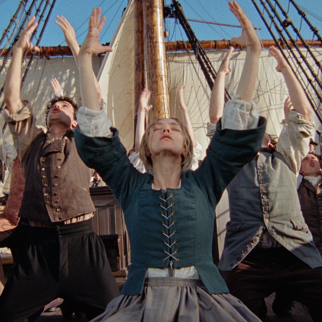 A group of people (center: Amanda Seyfried as Ann Lee) perch on their knees and raise their heads and hands to the sky. They wear 18th century clothing as they dance on the deck of a ship with a beige sail rising behind them, in THE TESTAMENT OF ANN LEE.