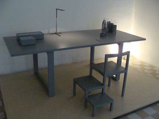 A grey rectangular table with 2 grey boxes on the left and bottled on the right and a matching grey chair (with stools attached) photographer on a platform against a white wall