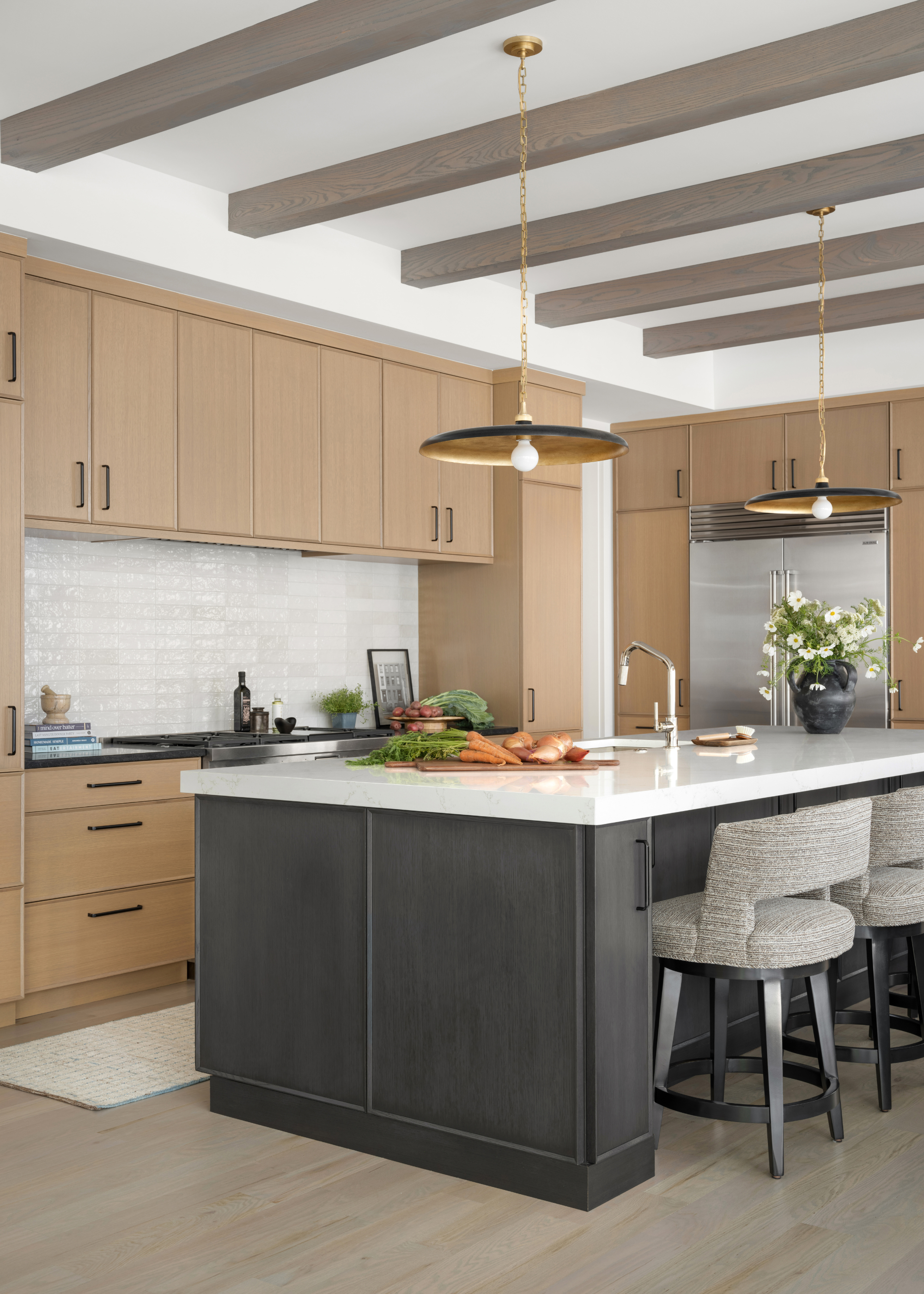A light wood kitchen with a black wood island, grey and black bar chairs and slim modern pendant lights hanging overhead