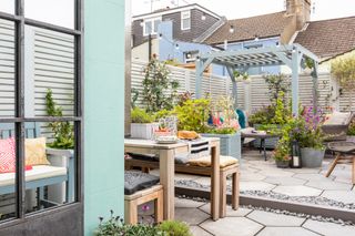 a paved garden with light blue painted raised planters and pergola, hexagonal paving, a grey mesh chair and footstool and light grey slatted fence
