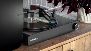 The Victrola Stream Onyx turntable pictured next to a Sonos speaker on one side and a plant on the other on a wooden shelf.