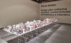 Side view of the show at the Prague Academy of Arts and Design featuring a long table filled with upright photographs in a room with white walls and grey flooring. There is black text on the back wall