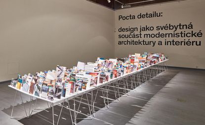 Side view of the show at the Prague Academy of Arts and Design featuring a long table filled with upright photographs in a room with white walls and grey flooring. There is black text on the back wall