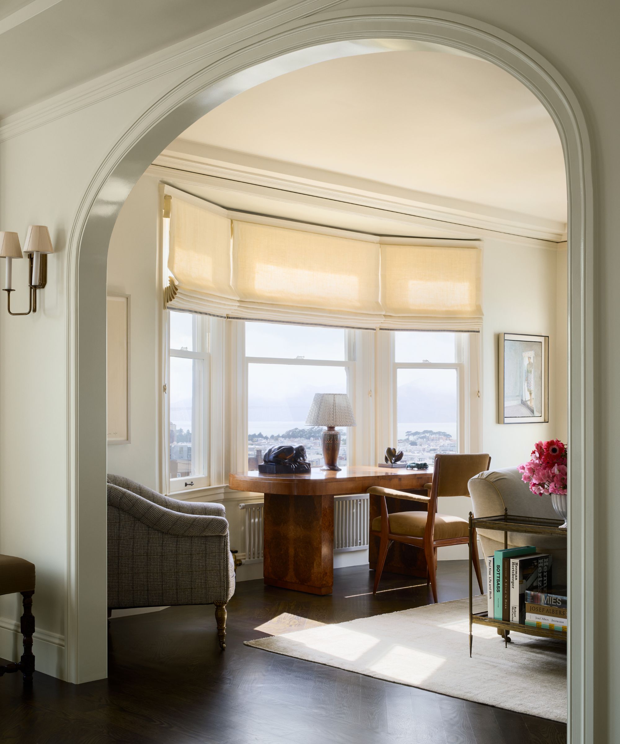 a soft green painted dining room looking through an arch to a cream living room with a bay window styled with a burl wood desk