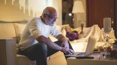 An older man looks at his laptop in his living room while his wife reads on the sofa in the background.