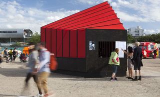 Alternative view of a black and red structure on stoney ground under a cloudy blue sky. There are buildings, an ice cream van and several people in the background