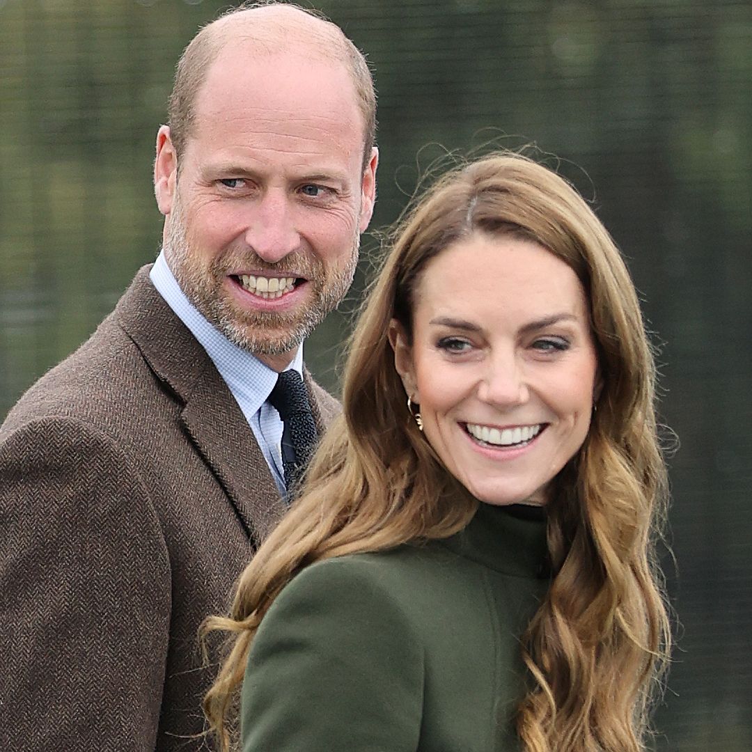 Prince William smiles while wearing a brown suit jacket and wife Kate Middleton wears a long green coat and has curly brown hair