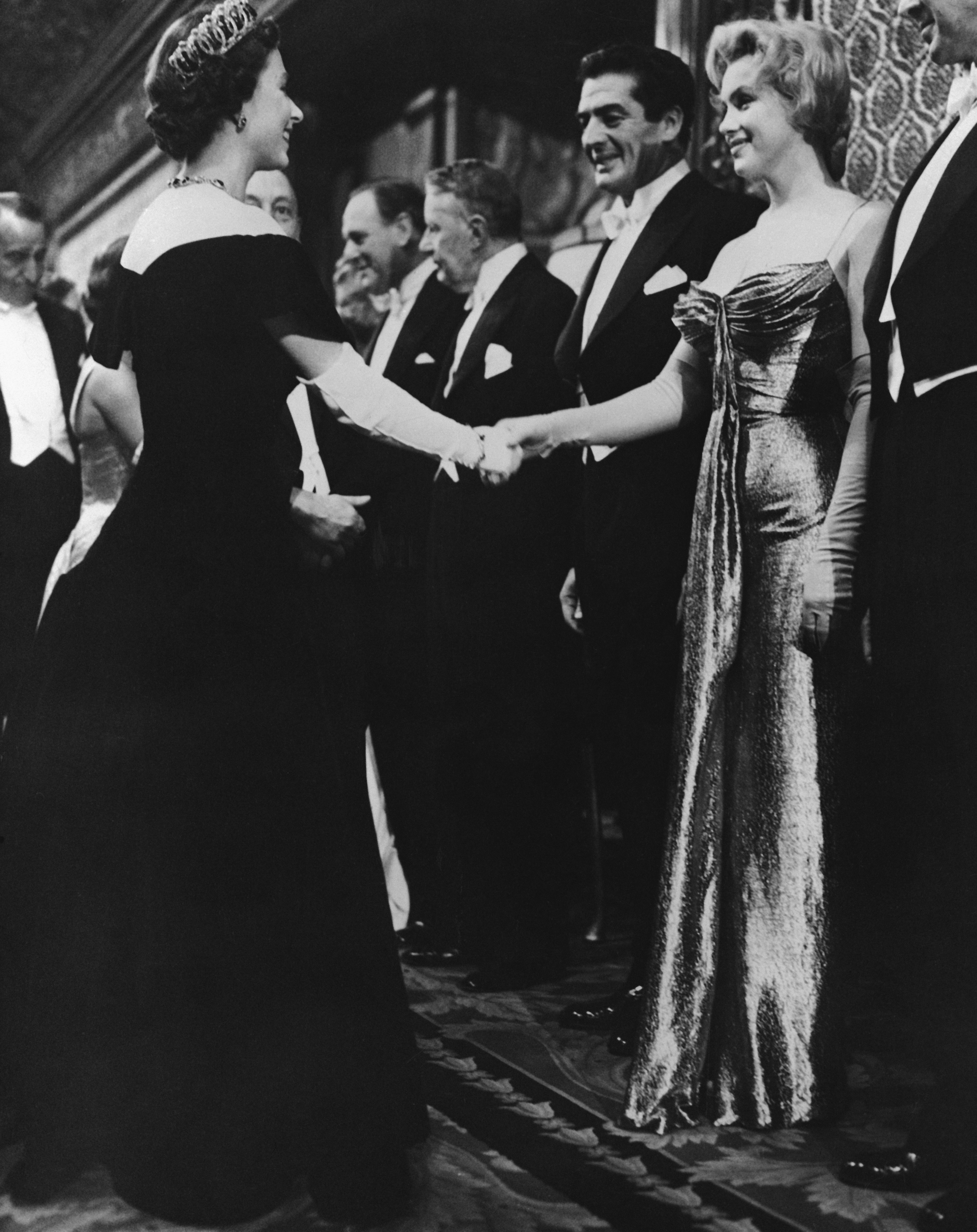 Queen Elizabeth II of England offers a gloved hand to Hollywood glamour girl Marilyn Monroe Miller during the Queen's visit with celebrities at the Royal Film Performance. Standing beside Marilyn is actor Victor Mature.