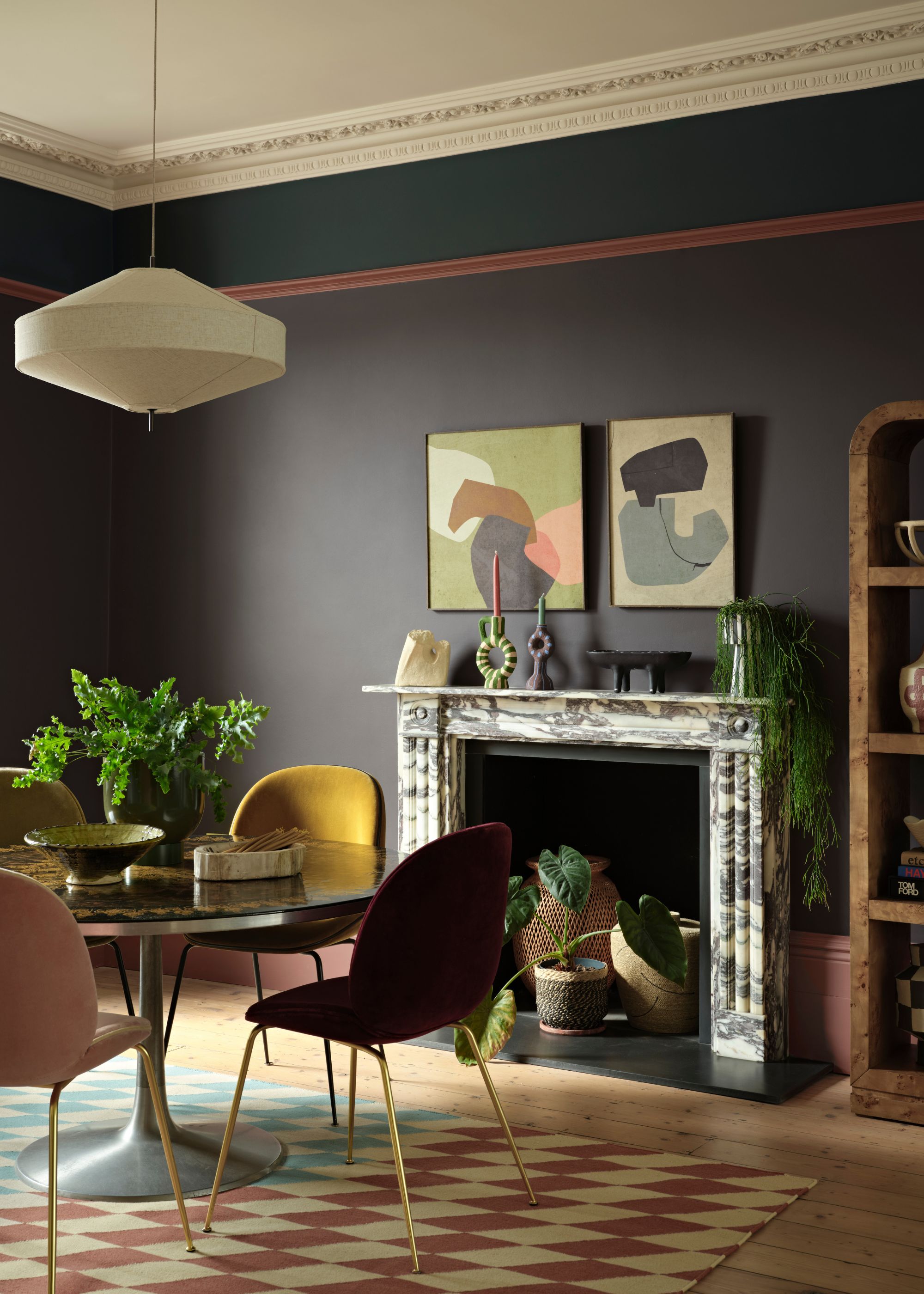 A dining room with dark brown-gray walls, a marble fireplace, a dining table, and colorful chairs.