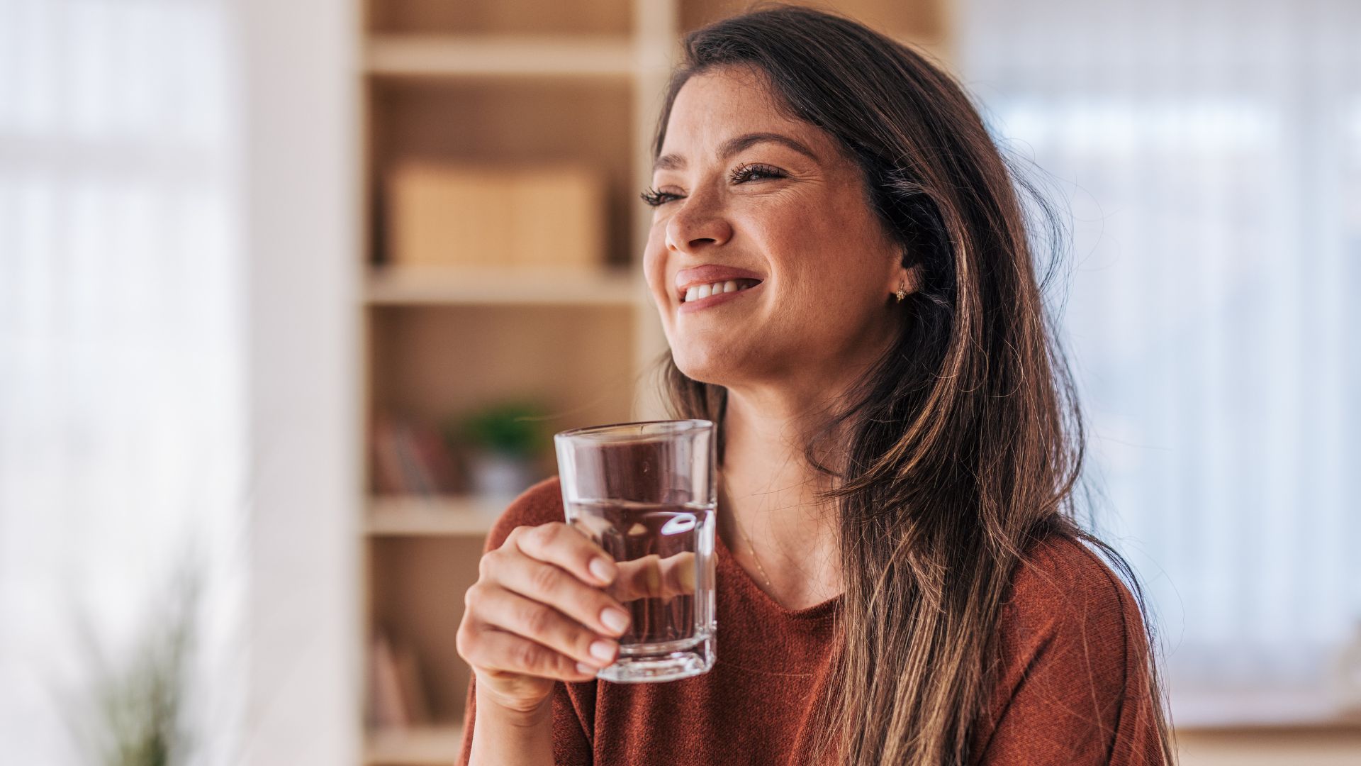 A woman standing in her home smiling as she holds a glass of water.