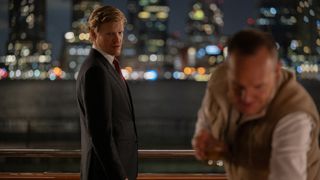 Jesse Plemons as Roger Carlson, dressed in a dark suit, white shirt and maroon tie, standing against a nighttime city backdrop, talking to an unidentified man.