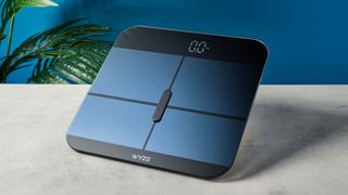 a sleek, clean-looking smart scale with tempered glass finish and a cross-shaped design on top of the glass, almost blue in the studio lighting, rests upon a table