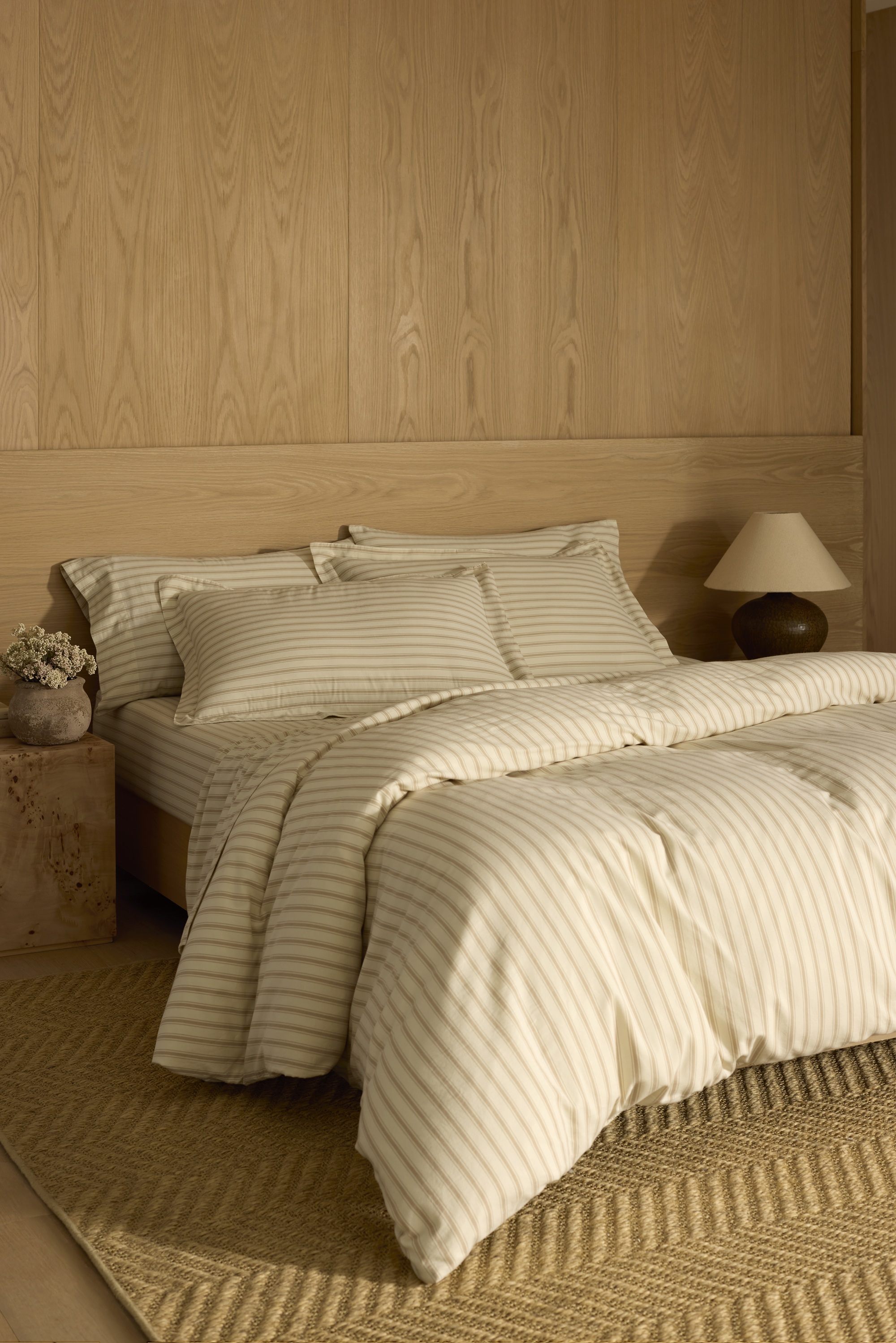 Cozy Earth soft wash cotton bedding in sandalwood stripe with timber wall and headboard, burl side table with vase of flowers, lamp, and jute rug