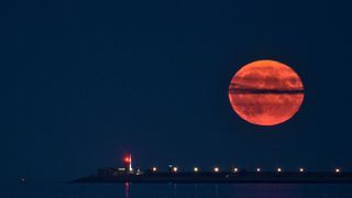 An orange full moon rises behind a thin cloud, against a dark blue sky, above a bridge lined with streetlights.