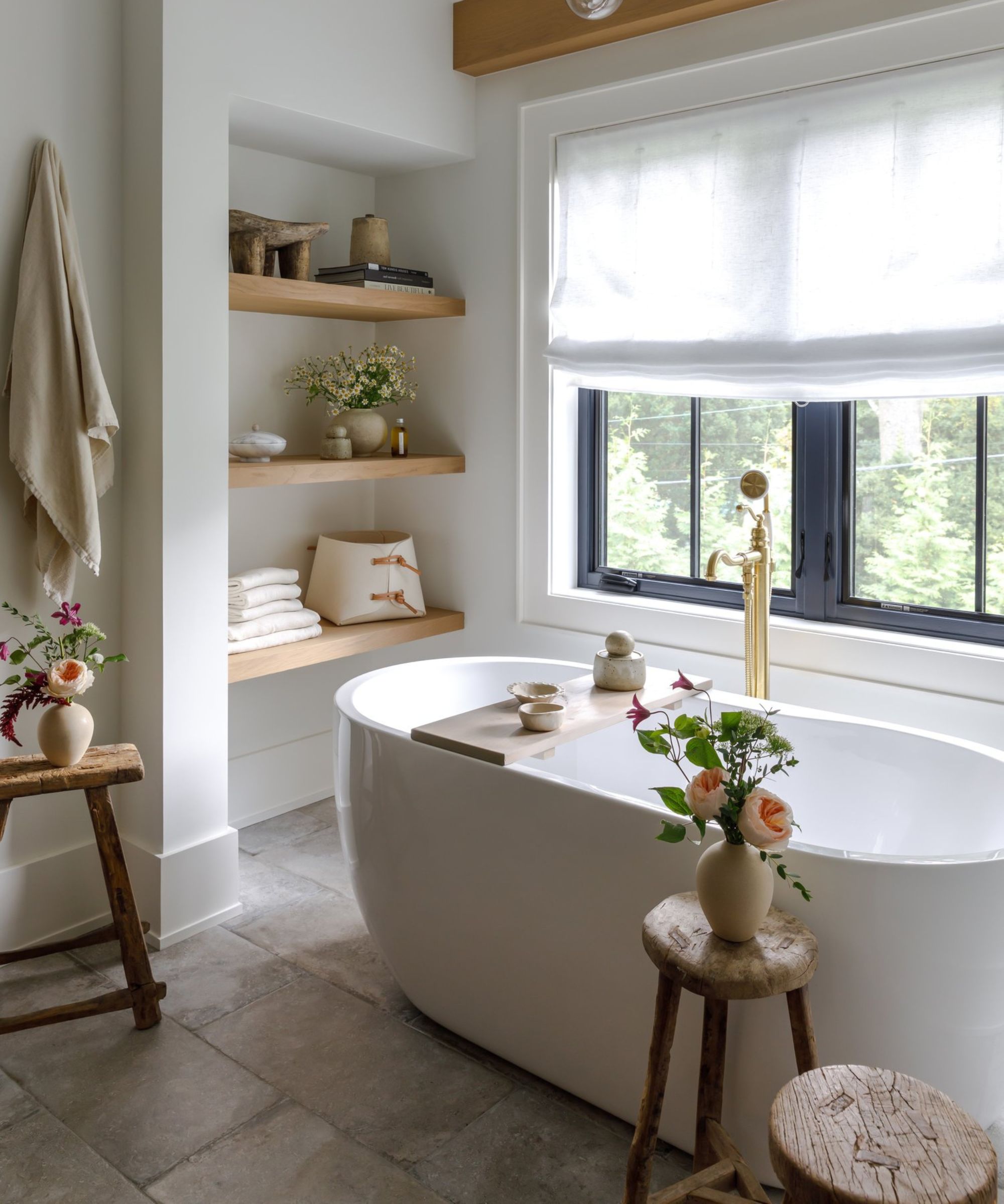modern rustic bathroom with stone floors, neutral walls, a modern freestanding bathtub with antique wooden stools styled with vases of flowers and wooden styled shelves