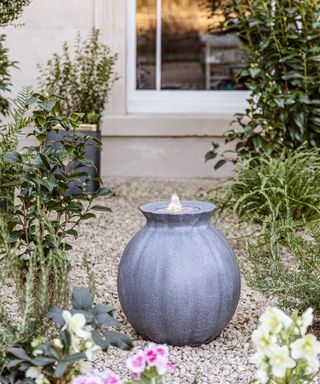 A grey round-shaped water feature stanging in a gravel garden around plants and flowers
