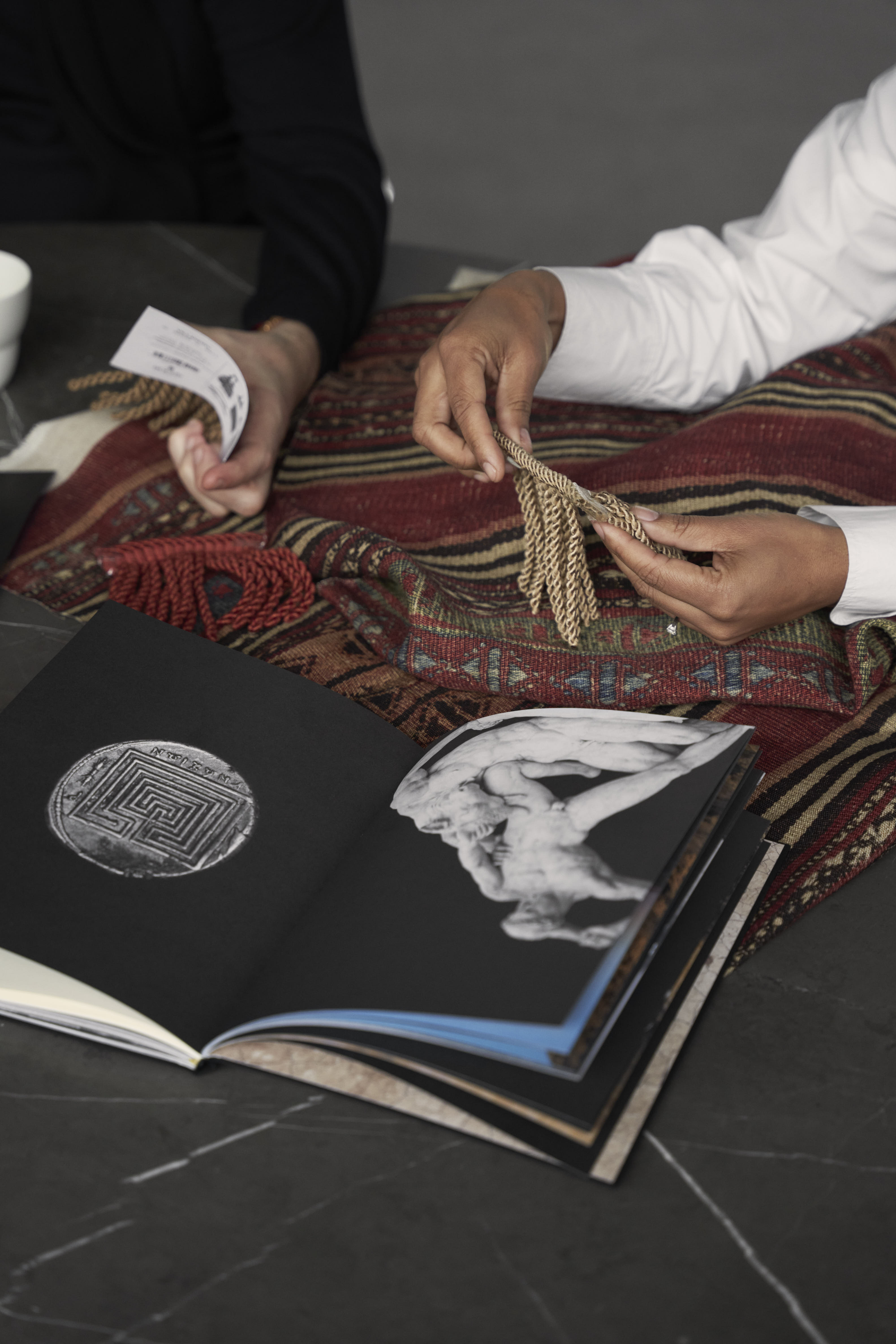 Two people sitting at a dark marble desk handle a series of notebooks with stunning imagery in them and rug textiles with a vibrant, earthy palette and geometrical motifs.