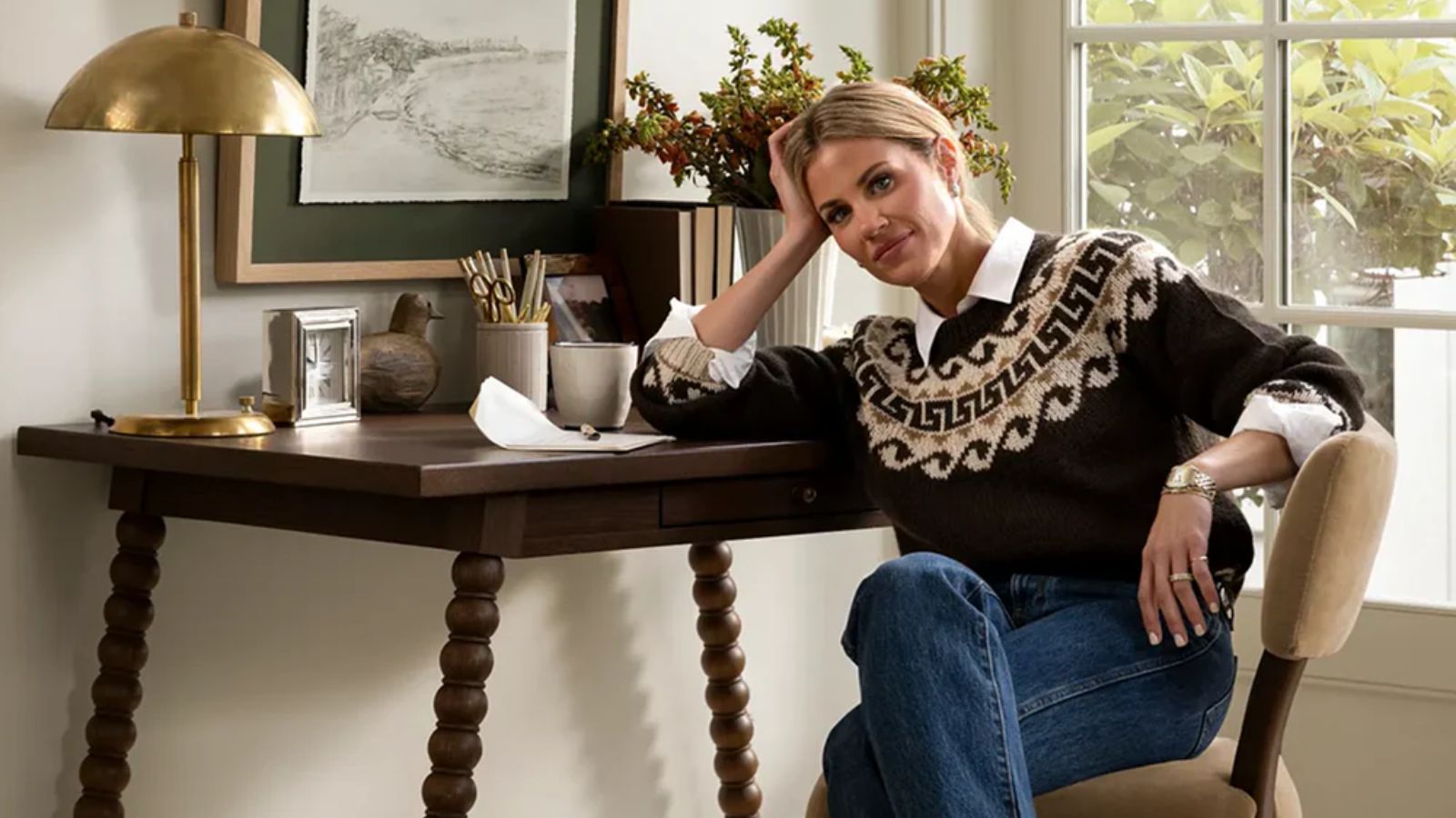 interior designer shea mcgee photographed for the mcgee and co fall collection 2025 sat by a bobbin dark wood desk on a neutral chair wearing jeans and a knitted jumper