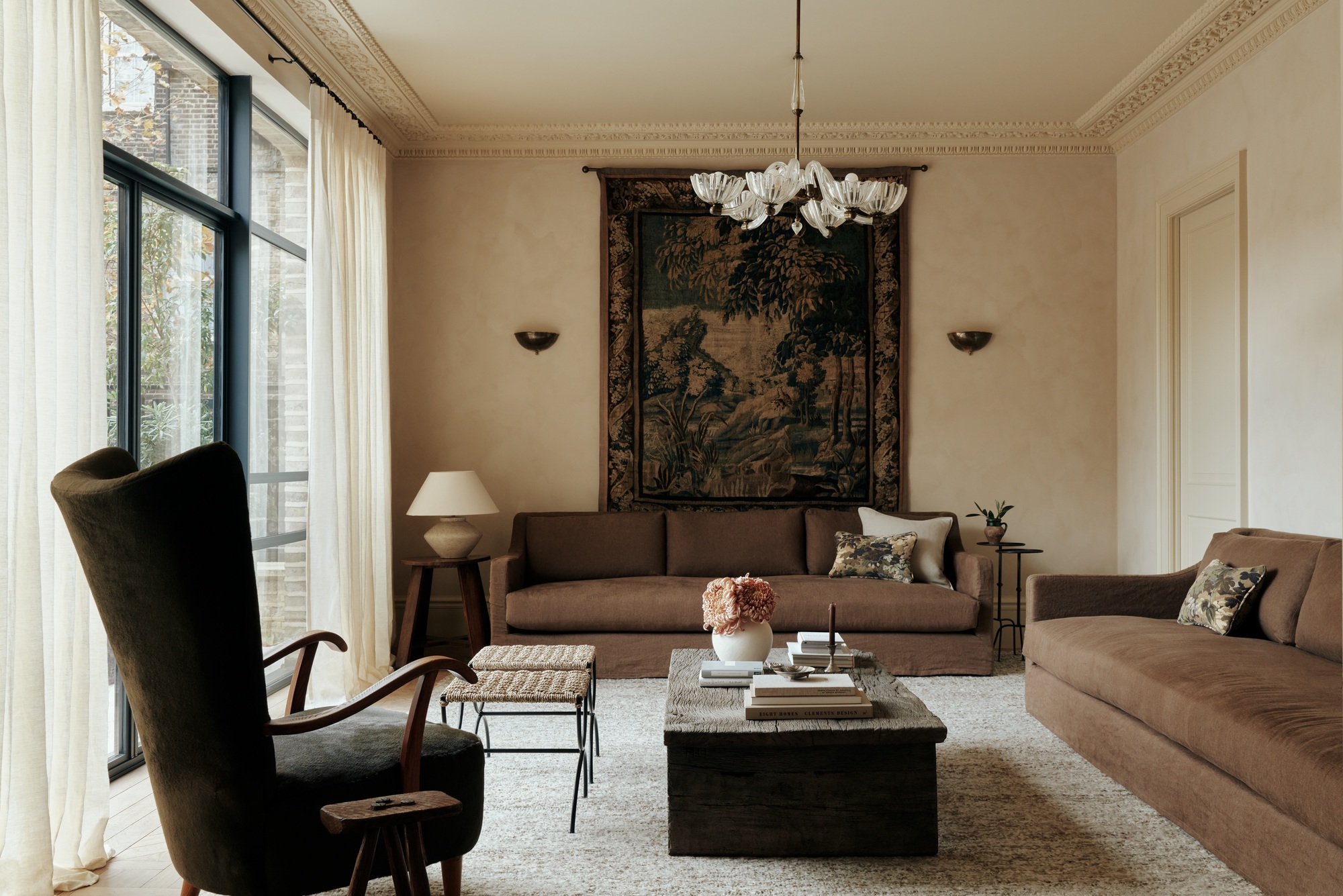 modern living room with wall tapestry, vintage ceiling light, brown sofas, timber coffee table styled with flowers and books, two rattan and steel stools, and a wing-backed armchair